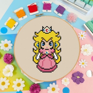Peut inclure: Un motif de point de croix représentant la Princesse Peach de la série de jeux vidéo Super Mario Bros. Le motif est dans un cercle de broderie et présente une image pixélisée de la Princesse Peach dans sa robe rose signature et ses cheveux jaunes.