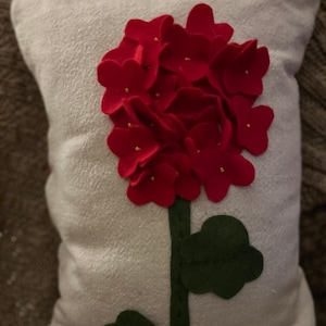 May include: A decorative pillow with a white background and a large, red felt flower design. The flower has a green stem and leaves. The pillow is square and appears to be made of a textured fabric.