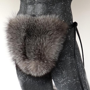 May include: A silver-grey fur accessory with a black leather strap. The fur is dense and plush, with variations in colour. The accessory is attached to a dark, textured mannequin torso.