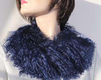 May include: A deep navy blue curly fur cowl scarf worn around the neck. The accessory features long, textured, and voluminous fibers that create a dense, shaggy appearance. The scarf is positioned over a light gray top, highlighting the dark color.