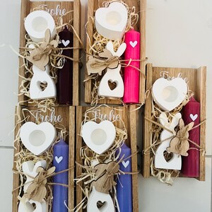 May include: Wooden candle holders with white heart-shaped candle holders, white bunny cutouts, and colored candles. The candles are purple, pink, and burgundy. The word "Frohe" is written on top of the wooden holders.