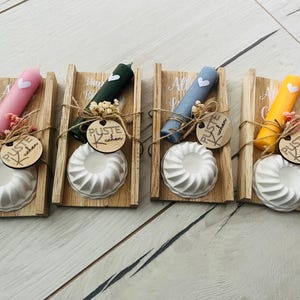 Puede incluir: Cuatro cajas de madera con un molde de cerámica blanca, una vela y una pequeña etiqueta de madera que dice "Puste Kuchen". Las cajas están decoradas con cintas y flores de diferentes colores.