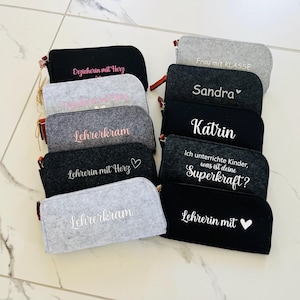 Puede incluir: Una colección de estuches de fieltro en varios colores, incluyendo negro, gris y antracita. Cada estuche presenta texto bordado en blanco o rosa, con frases como "Erzieherin mit Herz" y "Lehrerkram". Los estuches tienen cremalleras marrones.
