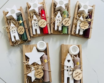 Puede incluir: Seis bandejas rectangulares de madera con conjuntos de decoración navideña. Cada set incluye un portavelas blanco en forma de estrella, un adorno de casa blanca, una vela cónica de color y una etiqueta de madera con el texto Frohe Weihnachten.