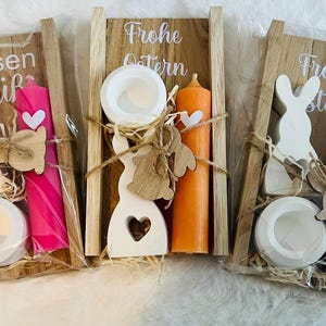 May include: Three Easter-themed gift sets. Each set includes a wooden bunny, a candle, and a small dish, all arranged in a wooden frame. The candles are pink, orange, and blue. The text "Frohe Ostern" and "Hasen gr&uuml;&szlig;" are visible.