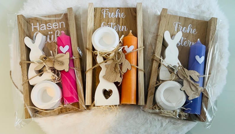 May include: Three Easter-themed gift sets. Each set includes a wooden bunny, a candle, and a small dish, all arranged in a wooden frame. The candles are pink, orange, and blue. The text "Frohe Ostern" and "Hasen gr&uuml;&szlig;" are visible.