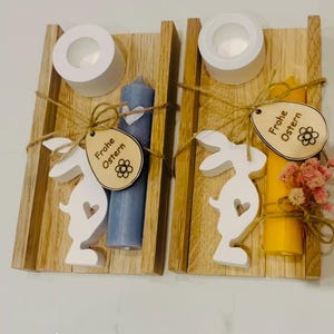 May include: Two wooden candle holders with candles, bunny cutouts, and tags that say "Frohe Ostern." One candle is blue, the other yellow. Each holder has a white tea light cup. The holders are tied with twine.
