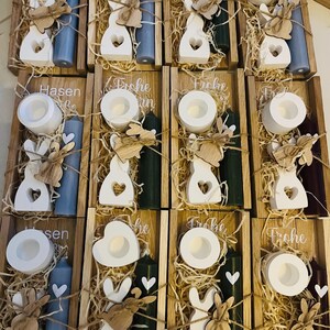 May include: Wooden trays filled with Easter decorations. Each tray contains a white candle holder, a white bunny cutout, a colored candle, and decorative straw. Some trays have the word "Frohe" or "Hasen" on them.