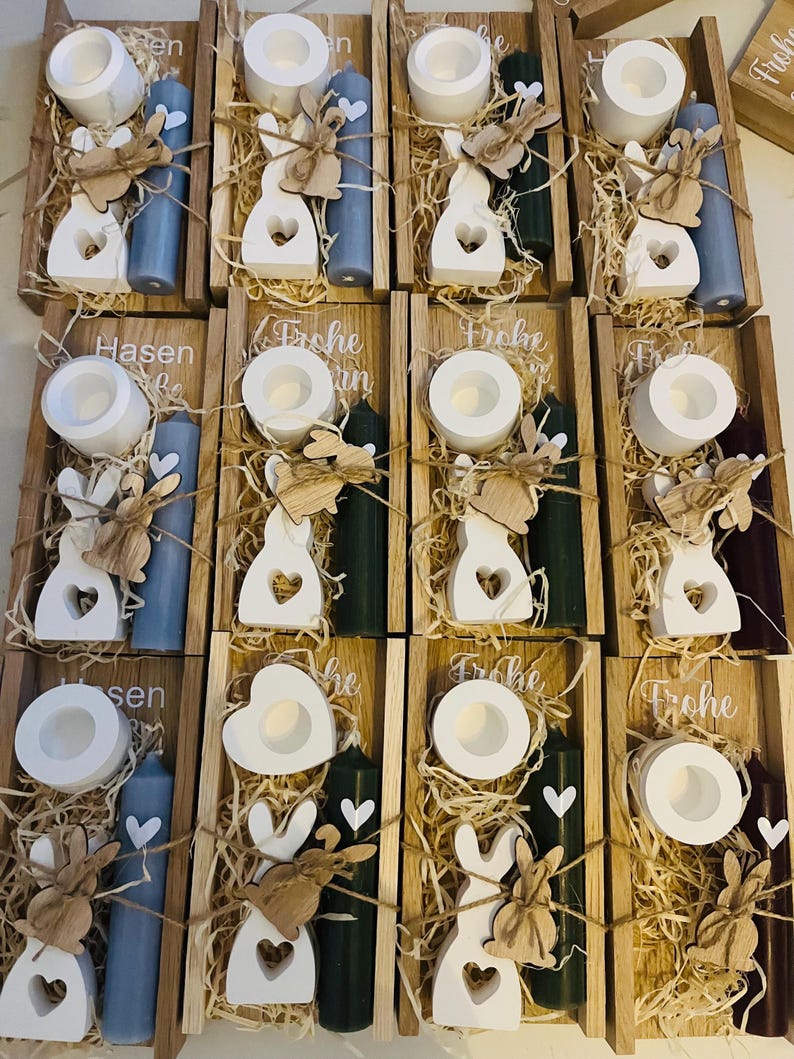 May include: Wooden trays filled with Easter decorations. Each tray contains a white candle holder, a white bunny cutout, a colored candle, and decorative straw. Some trays have the word "Frohe" or "Hasen" on them.