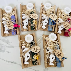 May include: Wooden trays with Easter-themed decorations. Each tray contains a candle, a white bunny cutout, decorative straw, and a wooden tag with the text "Frohe Ostern". The candles are in various colors, including purple, green, and blue.