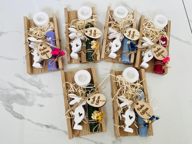 May include: Wooden trays with Easter-themed decorations. Each tray contains a candle, a white bunny cutout, decorative straw, and a wooden tag with the text "Frohe Ostern". The candles are in various colors, including purple, green, and blue.