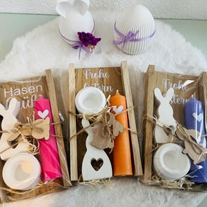 May include: Easter-themed decorative items. Includes three wooden trays with candles, bunny cutouts, and candle holders. Two white decorative eggs and a bunny-shaped decoration are also present. The trays have text that reads "Frohe Ostern" and "Hasen".