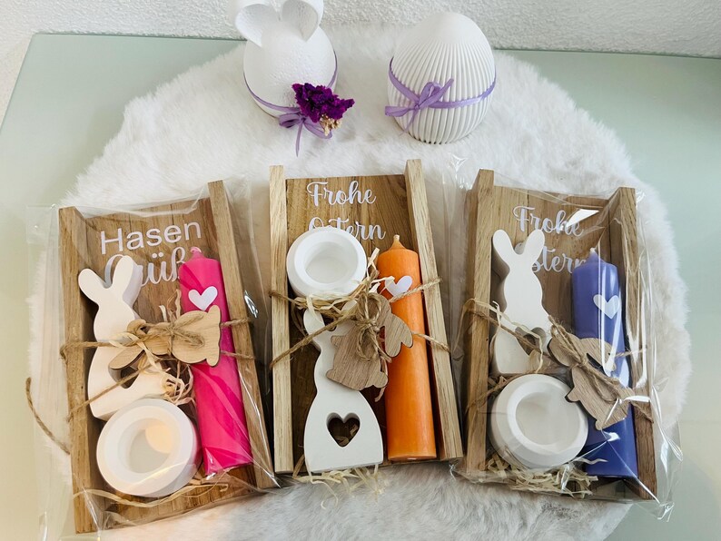 May include: Easter-themed decorative items. Includes three wooden trays with candles, bunny cutouts, and candle holders. Two white decorative eggs and a bunny-shaped decoration are also present. The trays have text that reads "Frohe Ostern" and "Hasen".