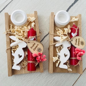 May include: Two wooden Easter decorations. Each features a small red candle, a white bunny cutout, and a wooden tag that says "Frohe Ostern". The decorations are in a wooden tray with decorative filler and small pink flowers.