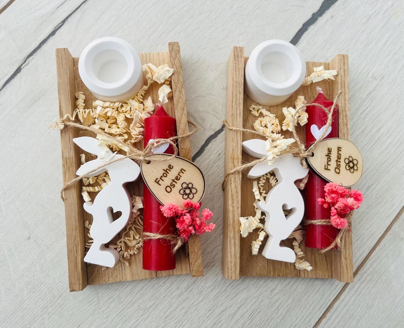 May include: Two wooden Easter decorations. Each features a small red candle, a white bunny cutout, and a wooden tag that says "Frohe Ostern". The decorations are in a wooden tray with decorative filler and small pink flowers.