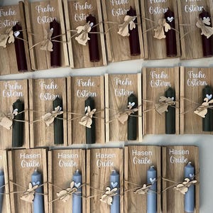 May include: A collection of Easter-themed candles. Each candle is affixed to a wooden plaque with a decorative wooden bunny and twine. The plaques feature the words "Frohe Ostern" and "Hasen Gr&uuml;&szlig;e" in white script, with candles in burgundy, green, and blue.