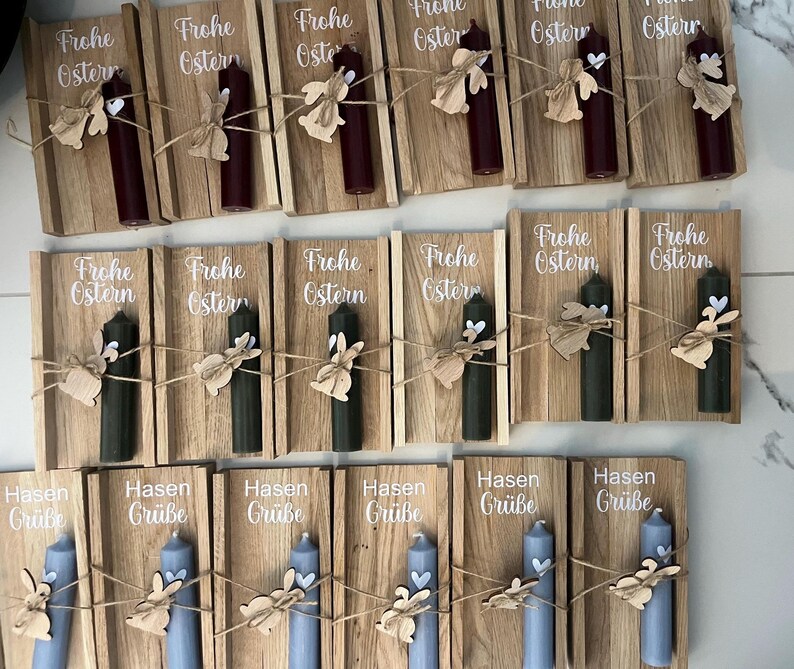 May include: A collection of Easter-themed candles. Each candle is affixed to a wooden plaque with a decorative wooden bunny and twine. The plaques feature the words "Frohe Ostern" and "Hasen Gr&uuml;&szlig;e" in white script, with candles in burgundy, green, and blue.