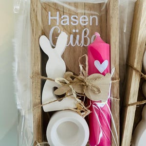 May include: A wooden Easter-themed gift set in a clear plastic wrap. The set includes a white bunny cutout, a pink candle with a white heart, a white ceramic egg holder, and a wooden sign with the text "Hasen Gr&uuml;&szlig;e".