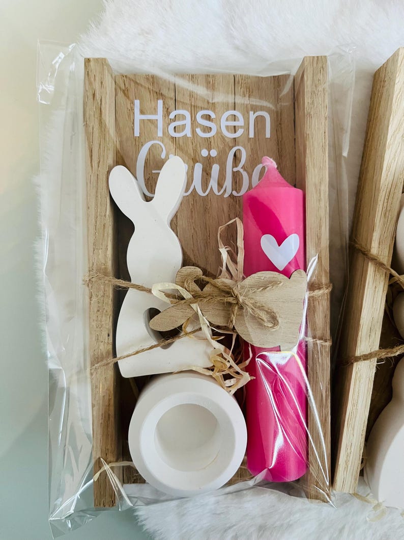 May include: A wooden Easter-themed gift set in a clear plastic wrap. The set includes a white bunny cutout, a pink candle with a white heart, a white ceramic egg holder, and a wooden sign with the text "Hasen Gr&uuml;&szlig;e".