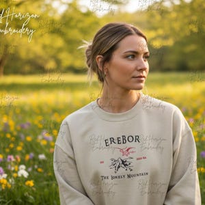 May include: A beige crewneck sweatshirt featuring a dark red and black embroidered graphic on the chest. The design includes the text EREBOR, a small red dragon, a mountain illustration, and the text The Lonely Mountain with EST 2001.