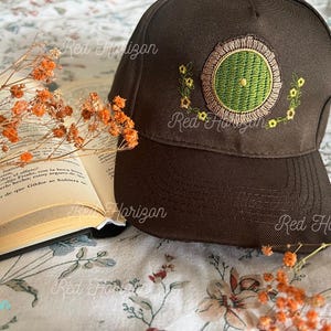Puede incluir: Gorra de béisbol marrón con un diseño bordado en verde y amarillo. El diseño es un emblema circular con un centro verde y un borde floral amarillo. La gorra está junto a un libro abierto con flores naranjas.