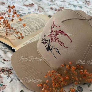 Puede incluir: Gorra de béisbol beige con un diseño bordado en rojo y negro. La gorra está colocada junto a un libro abierto y flores naranjas secas. Las palabras "Red Horizon" están impresas en la gorra.