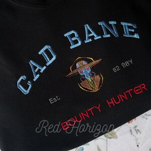 K&ouml;nnte beinhalten: Schwarzes Sweatshirt mit gesticktem Text und Grafik. Der blaue Text lautet "GAD BANE", mit einer Charakterillustration und "BOUNTY HUNTER" in Rot. Das Sweatshirt hat auch den Text "Est. 62 BBY".