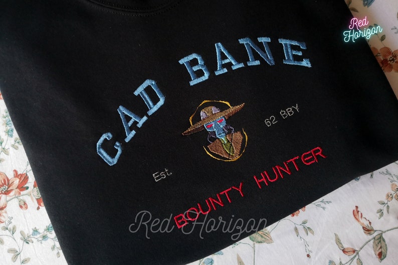 K&ouml;nnte beinhalten: Schwarzes Sweatshirt mit gesticktem Text und Grafik. Der blaue Text lautet "GAD BANE", mit einer Charakterillustration und "BOUNTY HUNTER" in Rot. Das Sweatshirt hat auch den Text "Est. 62 BBY".