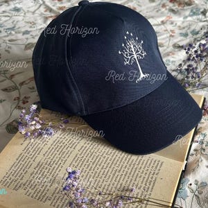 Op de afbeelding: Marineblauwe baseballpet met een wit boomontwerp. De pet ligt op een open boek met tekst en paarse bloemen. De pet heeft een gebogen klep en een knoop aan de bovenkant.