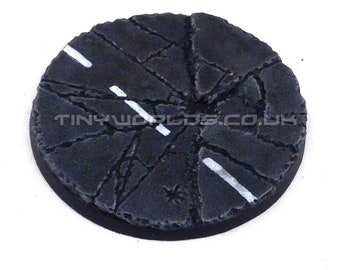 May include: A round, unpainted miniature base with a cracked asphalt road design. The base is dark gray with white painted road lines. The text "TINYWORLDS.CO.UK" is visible on the base. The text "Product Supplied Unpainted" is also visible.