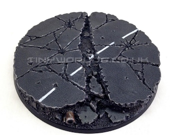 May include: A round, unpainted miniature base with a cracked asphalt design. The base features white road markings and a dark gray color scheme, with the text "Product Supplied Unpainted" at the bottom.