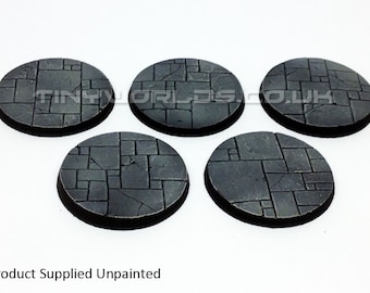 May include: Five unpainted, circular bases with a textured, stone-like pattern. The bases are dark gray with a black rim, and the surface is divided into irregular, rectangular shapes. The text "TINYWORLDS.CO.UK" is visible on the top.