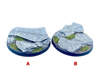 Può includere: Due basi circolari in miniatura etichettate A e B, caratterizzate da formazioni rocciose grigie testurizzate con macchie di erba verde. Le rocce presentano bordi frastagliati e dettagli realistici. Il testo Product Supplied Unpainted è visibile.