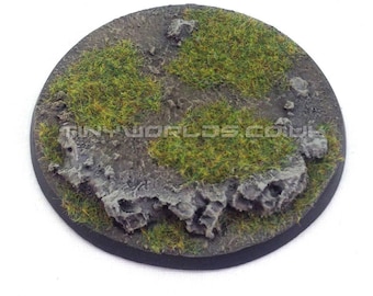 May include: A round, unpainted miniature base with a textured surface. The base features patches of green grass, rocky terrain, and a dark rim. The text "TINYWORLDS.COM.UK" is visible. The product is supplied unpainted.