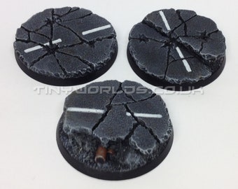 May include: Three unpainted, round miniature bases with a cracked asphalt design. Each base features a white painted road line. The bases are dark gray with a black rim. The text "Product Supplied Unpainted" is visible.
