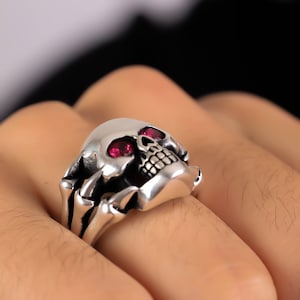 May include: A silver skull ring with red gemstone eyes. The skull is detailed with teeth and a bony band around the ring.