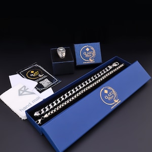 May include: A silver ring with a compass design in a blue box with the text "QART Jewelry" and "Hand Made Jewelry". A silver chain bracelet is in a larger blue box with the same text. A white cleaning cloth with the text "hand made" is also included.