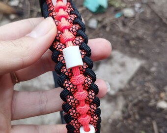 May include: A black and orange paracord bracelet with white beads. The bracelet is made with a woven pattern and has a black leather strap at the top.
