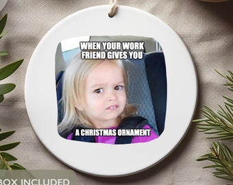 May include: White ceramic Christmas ornament with a picture of a young girl with a confused expression. The text on the ornament reads "When your work friend gives you a Christmas ornament."
