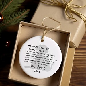 May include: White ceramic ornament with a silhouette of a dog and the text "veterinarian [noun] [vet] a person who has a big heart and a thoughtful mind, they have the strength to do what is right even when it hurts and will always have the deepest love for all creatures. Dr. Hart 2023"