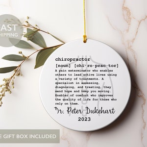 May include: White round ornament with a black and white definition of a chiropractor. The text includes the definition, "A pain exterminator who enables others to lead active lives using a variety of treatments." The ornament also includes the name "Dr. Peter Dukehart" and the year "2023".
