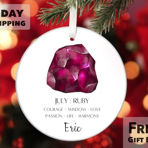May include: A white round ornament with a red ruby gemstone illustration. The ornament has the text "JULY: RUBY" and "COURAGE - WISDOM - LOVE" and "PASSION - LIFE - HARMONY". The name "Eric" is written at the bottom of the ornament.