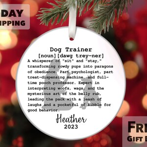 May include: White round Christmas ornament with a black silhouette of a dog trainer. The ornament has the text "Dog Trainer [noun] [dawg trey-ner] A whisperer of "sit" and "stay," transforming rowdy pups into paragons of obedience. Part psychologist, part treat-dispensing machine, and full-time pooch professor. Expert in interpreting woofs, wags, and the mysterious art of the belly rub, leading the pack with a leash of laughs and a pocketful of kibble for good behavior. Heather 2023" and "FREE GIFT BOX".