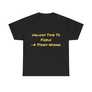 Może przedstawiać: Czarny t-shirt z żółtym napisem: "UNLUCKY TIME TO FIZZLE -A STORM WIZARD". Koszulka z okrągłym dekoltem w stylu casual. Tekst jest wyśrodkowany na przodzie koszulki.