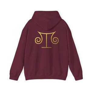 Peut inclure: Sweat-shirt à capuche bordeaux avec un symbole Sankofa doré au dos.