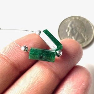 May include: Two rectangular, emerald green gemstone beads strung on a thin silver wire, with a silver bead on each end. A quarter coin is in the background. The gemstones are a deep, rich green color.