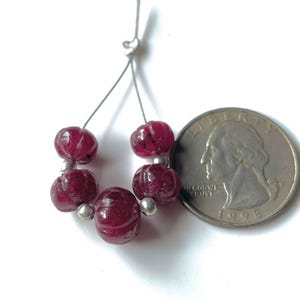May include: A strand of six dark red, carved gemstone beads with silver-colored spacers. A U.S. quarter coin is next to the beads for size comparison. The beads are strung on a thin, silver-colored wire.