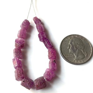 May include: A strand of rough-cut, vibrant pink ruby beads. The beads are strung on a thin wire, with a quarter coin next to the beads for size comparison. The beads have a natural, unpolished appearance.