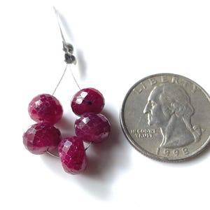 May include: A strand of faceted, deep red ruby beads. The beads are round and have a sparkling, multi-faceted surface. A US quarter is next to the beads for size comparison. The beads are strung on a thin, silver-colored wire.