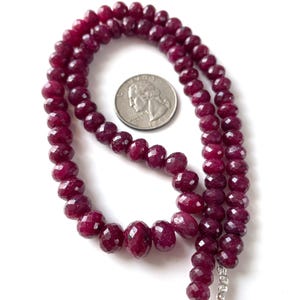 May include: A strand of faceted, deep red ruby beads. A U.S. quarter coin is placed near the beads for size comparison. The beads are strung together, with a tassel at the end. The background is white.